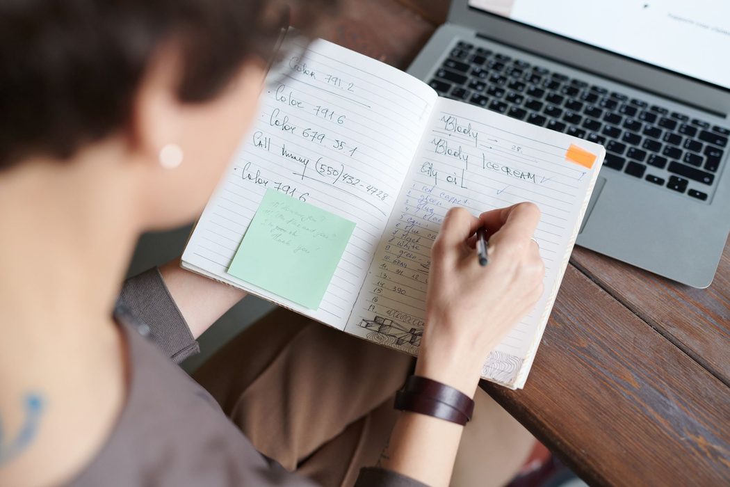 A writer taking notes at a desk beside an open laptop